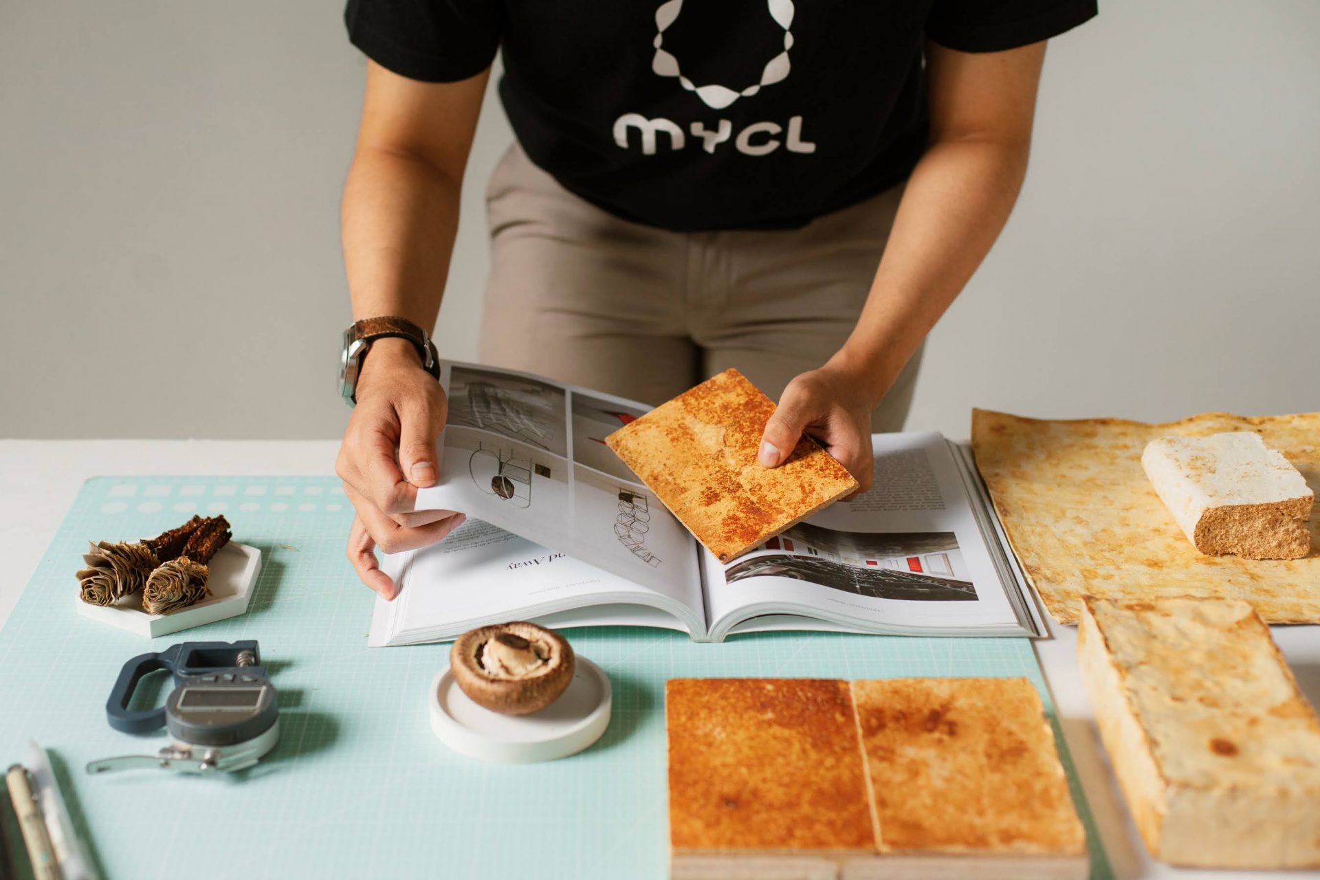 A person wearing a black MYCL shirt, handling several lmushroom leather sampleson a table.
