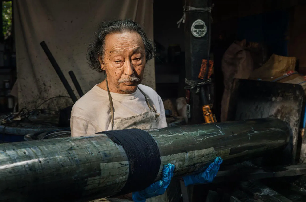 Kaei Hayakawa, a restoration artist of Arashi Shibori, holds a long wooden rod wrapped in fabric, carefully observing his work beside a vat of dye. In the dimly lit workshop, the seasoned craftsman stands in an apron, his expression focused and intent.