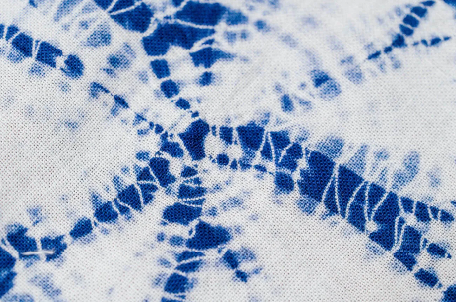 Close-up of a Narumi Shibori textile. Indigo-dyed, spiderweb-like patterns radiate across an unbleached white background, with each tied point creating delicate, ring-like contours that reveal the precision of the hand-binding technique.