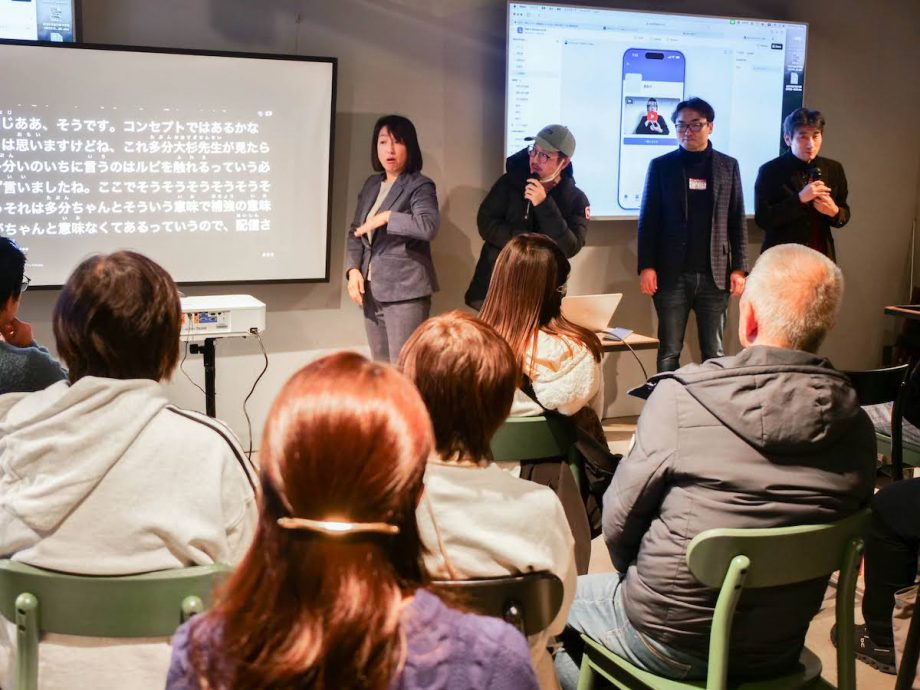 会場の参加者がモニタの前に立ちマイクを持って話している。左に手話通訳者、右には芝垣さんと奥田さん