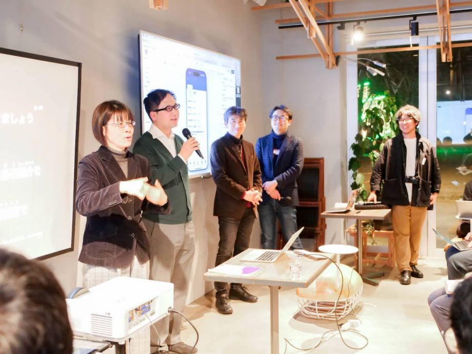 会場の参加者がモニタの前に立ちマイクを持って話している。左に手話通訳者、右には微笑む芝垣さんと奥田さん