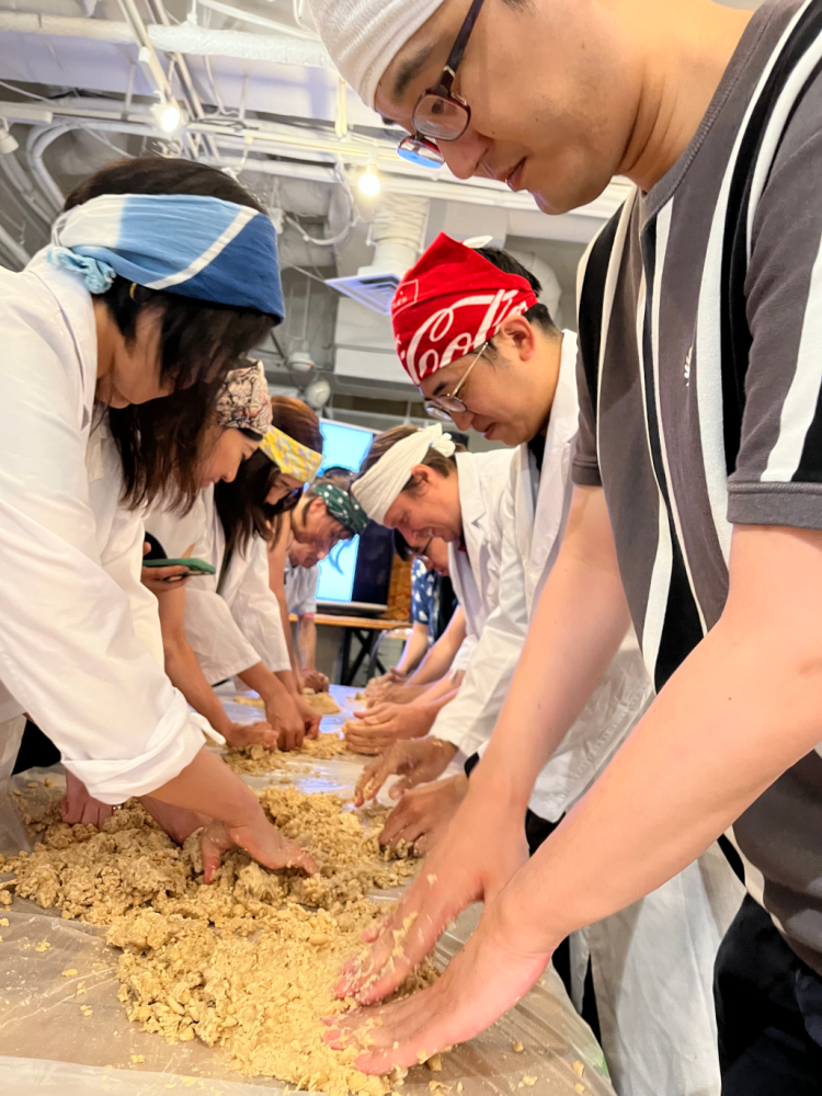 A photo of people standing alongside a table and squishing soybeans with their bare hands. They are wearing headbands; some are wearing lab coats.