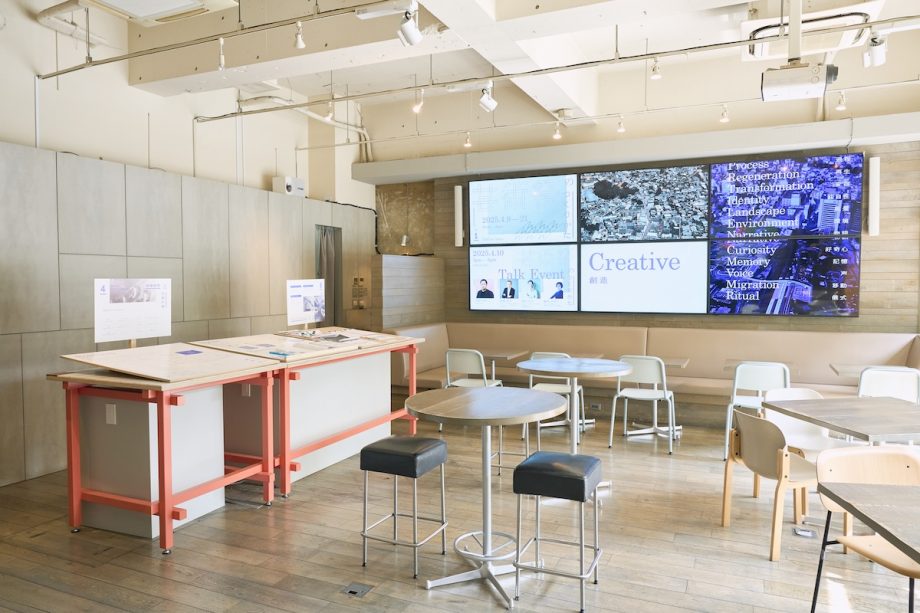 東京展の展示風景。カフェの座席の奥にある壁面の大型液晶ディスプレイでは、まちづくり・エリアマネジメントに対する芸術祭の意義を紹介する動画や、展覧会会期中に実施される関連イベントの情報が映し出されている。